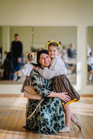 Valmiera, Latvia, April 1. 2024 - A smiling mother kneels to embrace her daughter in a dance studio, both looking at the camera, with mirrors reflecting the room.