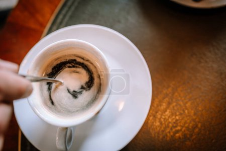 Valmiera, Lettland - 13. August 2023 - Ein Kopfschuss einer Hand, die eine halb leere Tasse Kaffee mit einem Löffel auf einer Untertasse auf einem strukturierten Tisch rührt..