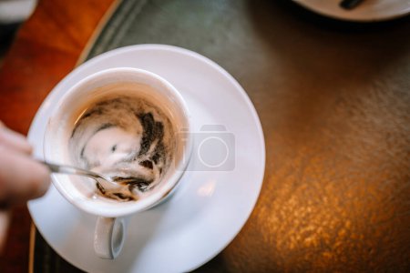 Valmiera, Lettland - 13. August 2023 - Ein Kopfschuss einer Hand, die eine halb leere Tasse Kaffee mit einem Löffel auf einer Untertasse auf einem strukturierten Tisch rührt..