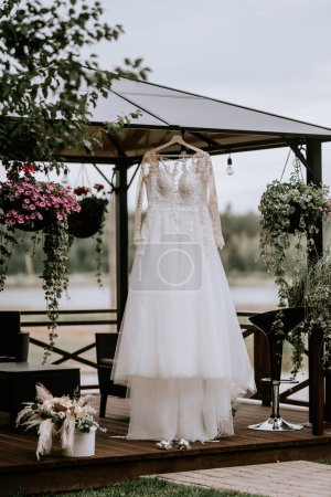 A white wedding dress with lace sleeves and bodice is displayed on a hanger outdoors under a gazebo with blooming flowers and a scenic pond in the background.