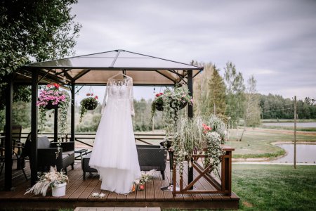 A white wedding dress with lace sleeves and bodice is displayed on a hanger outdoors under a gazebo with blooming flowers and a scenic pond in the background.