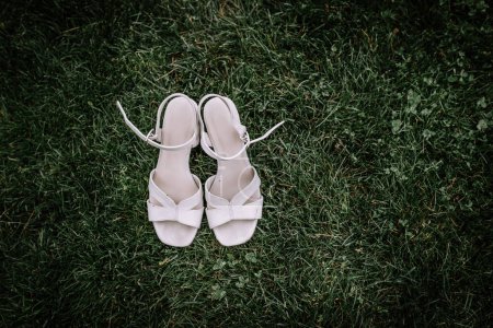 Valmiera, Lettland - 13. August 2023 - Ein Paar elegante weiße Sandalen mit Absatz werden auf einem sattgrünen Rasen platziert, was auf eine Hochzeit oder ein formelles Ereignis hindeutet..