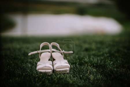 Elegant white bridal shoes on a grassy field with a blurred water body in the background, possibly near a wedding venue.
