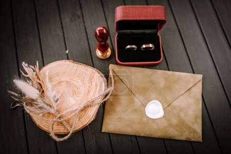 A wedding setup with golden wedding rings in a red box, a wax seal on a brown envelope, and a wooden ring dish adorned with dried flowers on a dark background.