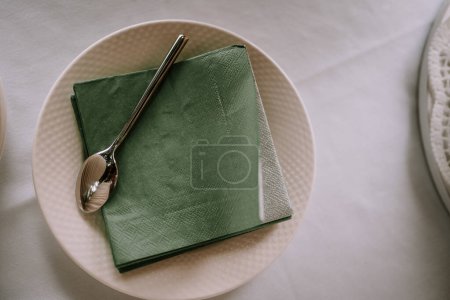 Valmiera, Latvia - Augist 13, 2023 - An overhead shot of a white ceramic plate with a textured edge, a green paper napkin, and a silver spoon on top.