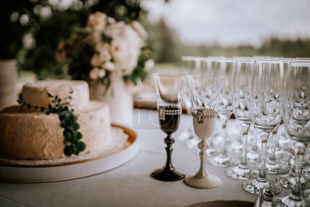 Close-up of a wedding cake on a table with two ornate glasses and a lineup of champagne flutes in the background, set for a celebration.