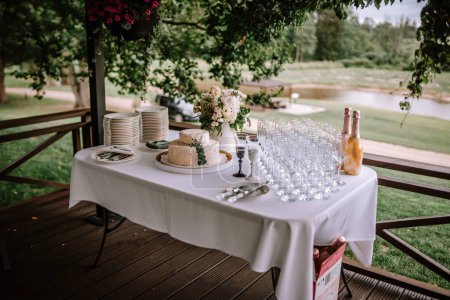 Valmiera, Latvia - Augist 13, 2023 - A wedding cake table set up outdoors with a two-tier cake, plates, champagne bottles, glasses, and a floral arrangement overlooking a serene pond.