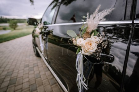 Ein Blumenschmuck mit weißen Rosen und Federn ist am Türgriff eines schwarzen Autos befestigt, wahrscheinlich Teil eines Hochzeitskonvois.