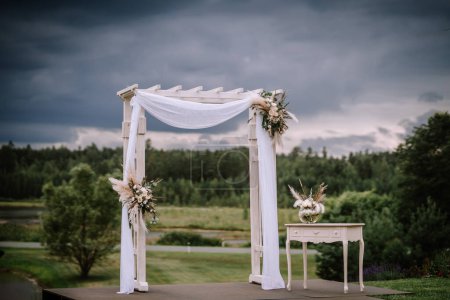 Photo for Valmiera, Latvia - Augist 13, 2023 - An elegant white wedding arch with draped fabric and floral arrangements stands under a moody sky, by a tranquil pond and green landscape. - Royalty Free Image