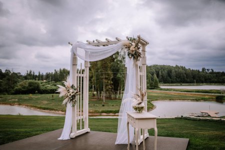 Valmiera, Latvia - Augist 13, 2023 - An elegant outdoor wedding arch adorned with white fabric and floral arrangements, set against a cloudy sky and a calm lake in the background.