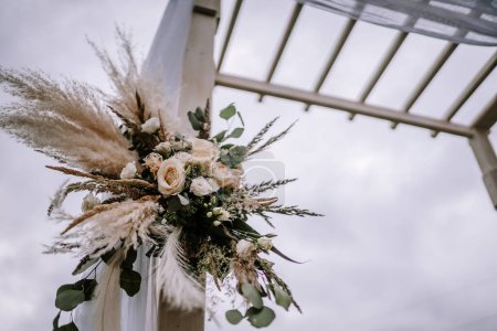 Nahaufnahme eines floralen Arrangements auf einem Hochzeitsbogen, mit einer Mischung aus Pampasgras, Rosen und Grün vor einer weißen Struktur und Himmelshintergrund.