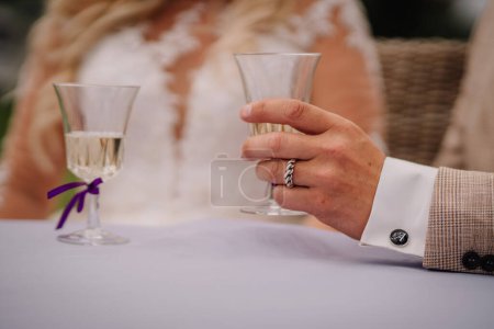 Photo for Valmiera, Latvia - Augist 13, 2023 - Close-up of a bride and groom's hands holding champagne glasses at a wedding, showcasing rings, elegant attire, and a hint of a floral bouquet. - Royalty Free Image