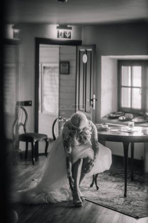Valmiera, Latvia - Augist 13, 2023 - A bride in a lace wedding dress is sitting down, fastening her sandal. The photo is monochrome, capturing a tranquil moment amidst wedding preparations.