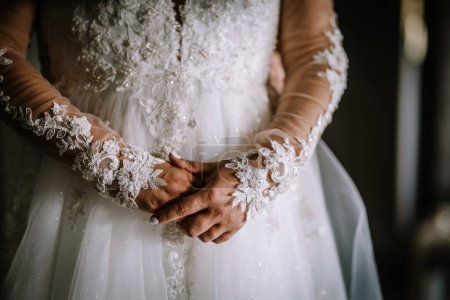 Valmiera, Lettonie - 13 août 2023 - Les mains jointes d'une mariée sont montrées avec des manches en dentelle détaillées et le corsage de sa robe de mariée, soulignant les motifs complexes et le travail de perles.