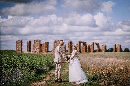 Foto de Una novia y un novio se toman de la mano en un campo con ruinas antiguas y un cielo vibrante arriba, que transmite un sentido del romance y la historia intemporales. - Imagen libre de derechos