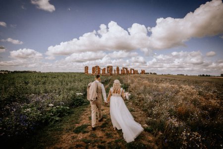 Photo for A couple walks hand in hand towards ancient ruins in a wildflower meadow under a dramatic sky, evoking a journey and adventure together. - Royalty Free Image