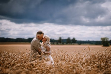 Photo for A bride and groom embrace and kiss in a wheat field, under a dramatic sky with looming clouds. - Royalty Free Image