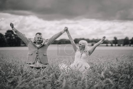 Foto de Una novia y un novio jubilosos toman de la mano y levantan sus brazos en triunfo en medio de un campo de hierba alta, con un cielo tormentoso por encima. - Imagen libre de derechos