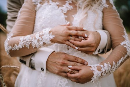 A close-up of a bride and groom's hands as they tenderly hold each other, showcasing their wedding rings and the intricate lace details of the bride's dress sleeve.