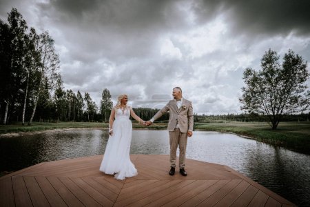 Foto de Valmiera, Letonia - 13 de agosto de 2023 - Una novia y un novio se toman de la mano en un muelle de madera que se extiende en un lago tranquilo, rodeado de un exuberante bosque y cielo nublado. - Imagen libre de derechos