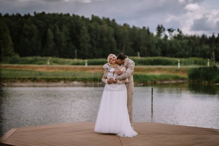 Valmiera, Latvia - Augist 13, 2023 - A bride and groom embrace on a wooden pier by the lake, the groom kisses the bride's cheek, both dressed in wedding attire, with a forest in the background.