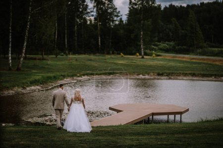 Valmiera, Latvia - Augist 13, 2023 - A bride and groom, seen from behind, walk hand in hand towards a serene lake with a modern wooden dock, surrounded by a lush green landscape.