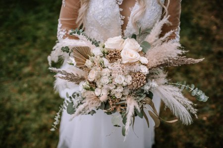 Valmiera, Lettland - 13. August 2023 - Eine Braut hält einen großen, kunstvollen Strauß mit Rosen, Pampasgras und verschiedenem Grün, der sich auf die Blumen konzentriert und im Hintergrund das Spitzenkleid der Braut zeigt..