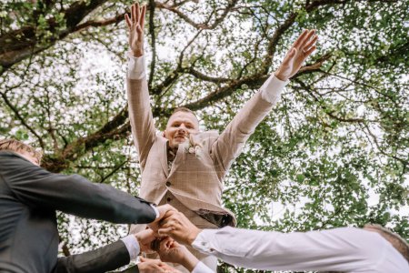 Valmiera, Latvia - Augist 13, 2023 - A groom is lifted into the air by his friends under a canopy of tree branches, joyfully celebrating with arms raised.