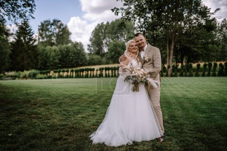 Valmiera, Latvia - Augist 13, 2023 - A bride in a white dress and a groom in a beige suit are embracing on a lush lawn, smiling with trees in the background.