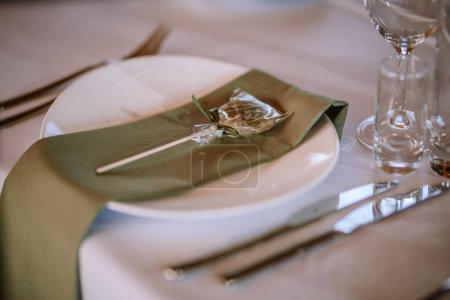 Close-up of a wedding table setting with a white plate, green cloth napkin, a decorative cookie wrapped in cellophane, and wooden cutlery, with clear glasses in the background.