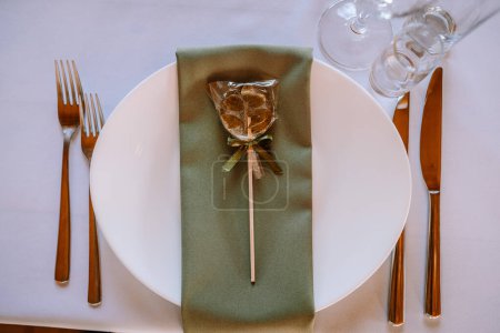 Valmiera, Letonia - 13 de agosto de 2023 - Elegante plato de comedor con una servilleta verde doblada, cubiertos de madera y una galleta decorativa envuelta en un embalaje transparente atado con una cinta de oro.