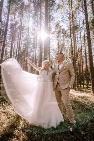 Foto de Valmiera, Letonia - 13 de agosto de 2023 - Una novia radiante con un vestido que fluye y su novio de pie en un bosque de pinos iluminado por el sol, su velo levantado por la brisa mientras se miran el uno al otro. - Imagen libre de derechos
