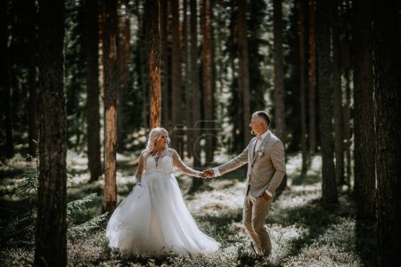 Photo for Valmiera, Latvia - Augist 13, 2023 - A bride and groom walk hand in hand through a pine forest, sunlight filtering through the trees, highlighting their joyful expressions. - Royalty Free Image