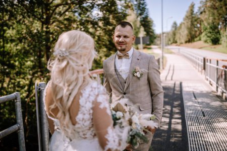 Foto de Valmiera, Letonia - 13 de agosto de 2023 - Un novio mira a la novia, que se aleja de la cámara, en una acera iluminada por el sol con vegetación en el fondo. - Imagen libre de derechos