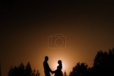 Photo for Valmiera, Latvia - August 13, 2023 - A silhouetted couple stands against a warm glowing sky, holding hands, with trees framing the scene and the sun setting directly behind them. - Royalty Free Image