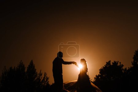 Foto de Valmiera, Letonia - 13 de agosto de 2023 - una novia y un novio se siluetean contra un cielo naranja dramático al atardecer, tocando amorosamente la frente con el sol entre ellos. - Imagen libre de derechos