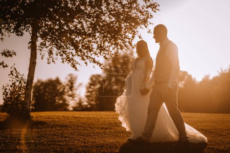 Photo for Valmiera, Latvia - Augist 13, 2023 - Silhouettes of a bride and groom holding hands against a sunset, with trees and a soft-focus background. - Royalty Free Image