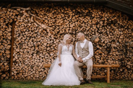 Valmiera, Latvia - Augist 13, 2023 - A bride and groom are sitting on a bench in front of a stacked woodpile, looking at each other affectionately.