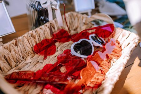 Valmiera, Latvia - Augist 13, 2023 - White heart-shaped sunglasses lie among red feathers in a wicker basket.