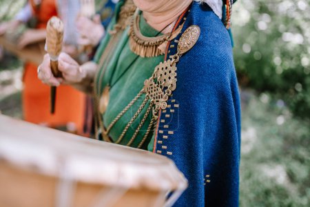 Valmiera, Latvia - July 14, 2023 - A close-up of a person in a blue shawl and green dress, adorned with traditional Latvian folk jewelry, playing a percussion instrument.