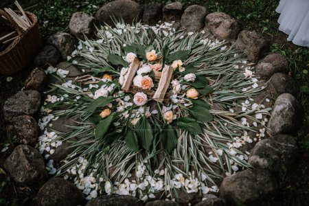 Valmiera, Letonia - 14 de julio de 2023 - Un arreglo floral popular letón ceremonial en el suelo, con flores blancas y de melocotón, hojas verdes y paja, rodeado de rocas.