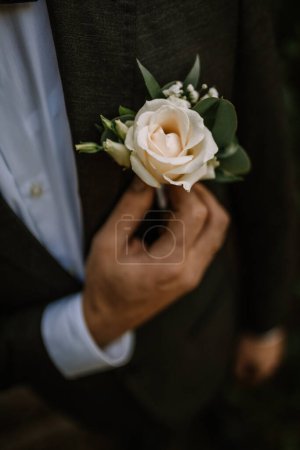 Valmiera, Latvia - July 14, 2023 - A close-up of a groom's hand adjusting a boutonniere with a white rose on the lapel of his dark suit.