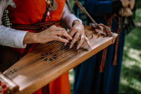 Valmiera, Lettland - 14. Juli 2023 - Hände spielen eine Kokle, ein traditionelles lettisches Saiteninstrument, im Hintergrund verschwimmen Musiker in Trachten.
