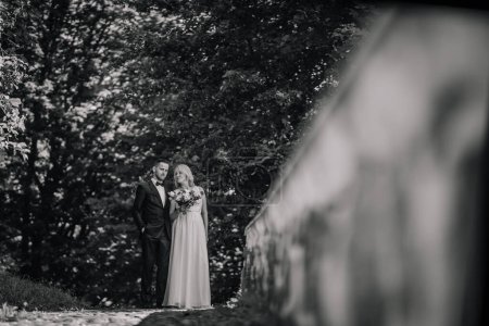 Valmiera, Lettonie - 14 juillet 2023 - Une photo en noir et blanc d'un marié en costume et d'une mariée tenant un bouquet debout ensemble dans un cadre extérieur serein et feuillu, évoquant un sentiment de romance et d "élégance.