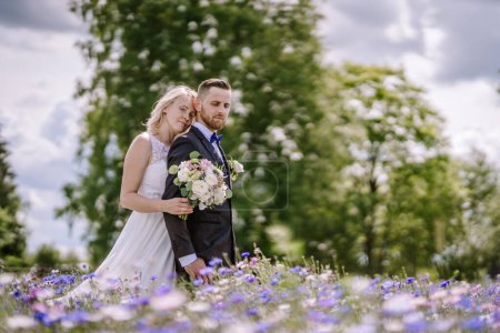 Foto de Valmiera, Letonia - 14 de julio de 2023 - Un abrazo de novia y novio en un campo, rodeado de flores silvestres con árboles y un cielo nublado en el fondo. - Imagen libre de derechos