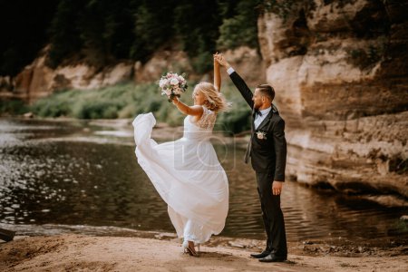 Valmiera, Letonia - 14 de julio de 2023 - Una novia y un novio alegres están de pie junto a un río, la novia sostiene su ramo en alto, su vestido ondeando, y el novio a su lado con el brazo levantado.