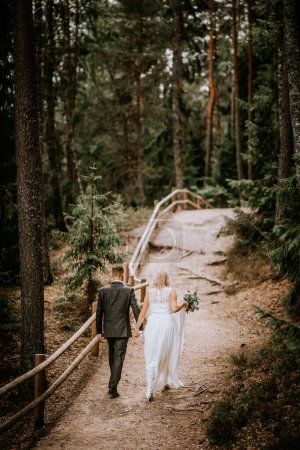 Photo for Valmiera, Latvia - July 14, 2023 - A bride and groom walk hand in hand down a forest path, with the groom leading and the bride following, holding her dress and a bouquet, in a serene woodland setting. - Royalty Free Image
