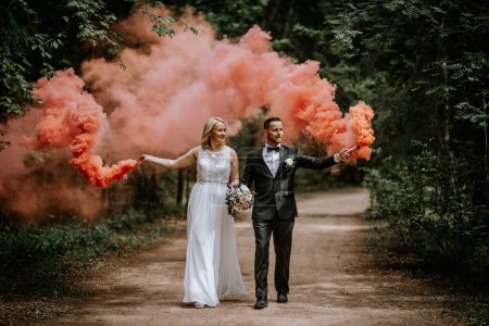 Photo for Valmiera, Latvia - July 14, 2023 - A bride and groom walk hand in hand in a forest, each holding a smoke flare emitting a dense, pink smoke, adding a whimsical touch to the scene. - Royalty Free Image