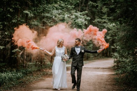 Foto de Valmiera, Letonia - 14 de julio de 2023 - Una novia y un novio caminan de la mano en un bosque, cada uno sosteniendo una llamarada de humo que emite un denso humo rosa, agregando un toque caprichoso a la escena.. - Imagen libre de derechos