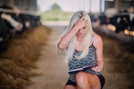 Téléchargez les photos : Valmiera, Lettonie - 17 août 2024 - Une femme avec une expression concernée est accroupie à lire un livre dans une grange avec des vaches en arrière-plan. - en image libre de droit